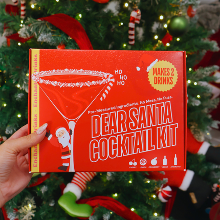 A hand holds a red box labeled 'Dear Santa Cocktail Kit' in front of a decorated Christmas tree with lights, ornaments, and stockings.