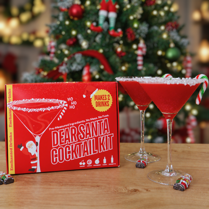 An Enchanted Drinks 'Dear Santa Cocktail Kit' box sits on a wooden table next to two martini glasses filled with shimmering red cocktails and candy cane garnishes, with a blurred decorated Christmas tree in the background.