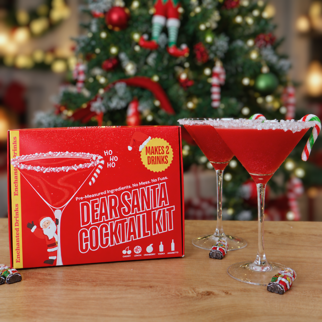An Enchanted Drinks 'Dear Santa Cocktail Kit' box sits on a wooden table next to two martini glasses filled with shimmering red cocktails and candy cane garnishes, with a blurred decorated Christmas tree in the background.