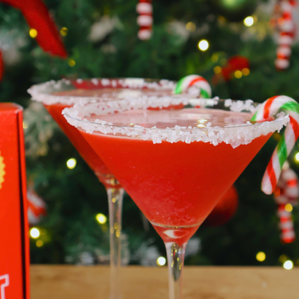 Two festive red Santa cocktails in martini glasses with sugar rims, garnished with candy canes, set against a blurred Christmas tree background with warm lights.